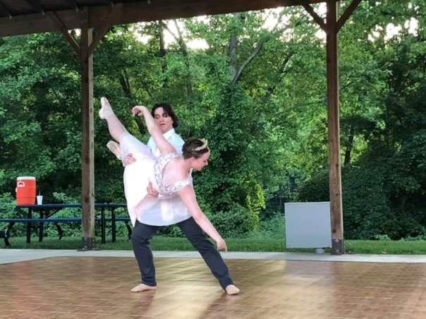The author holding his partner in a fish lift on an outdoor stage with greenery behind them and an orange beverage cooler to the viewer's left.