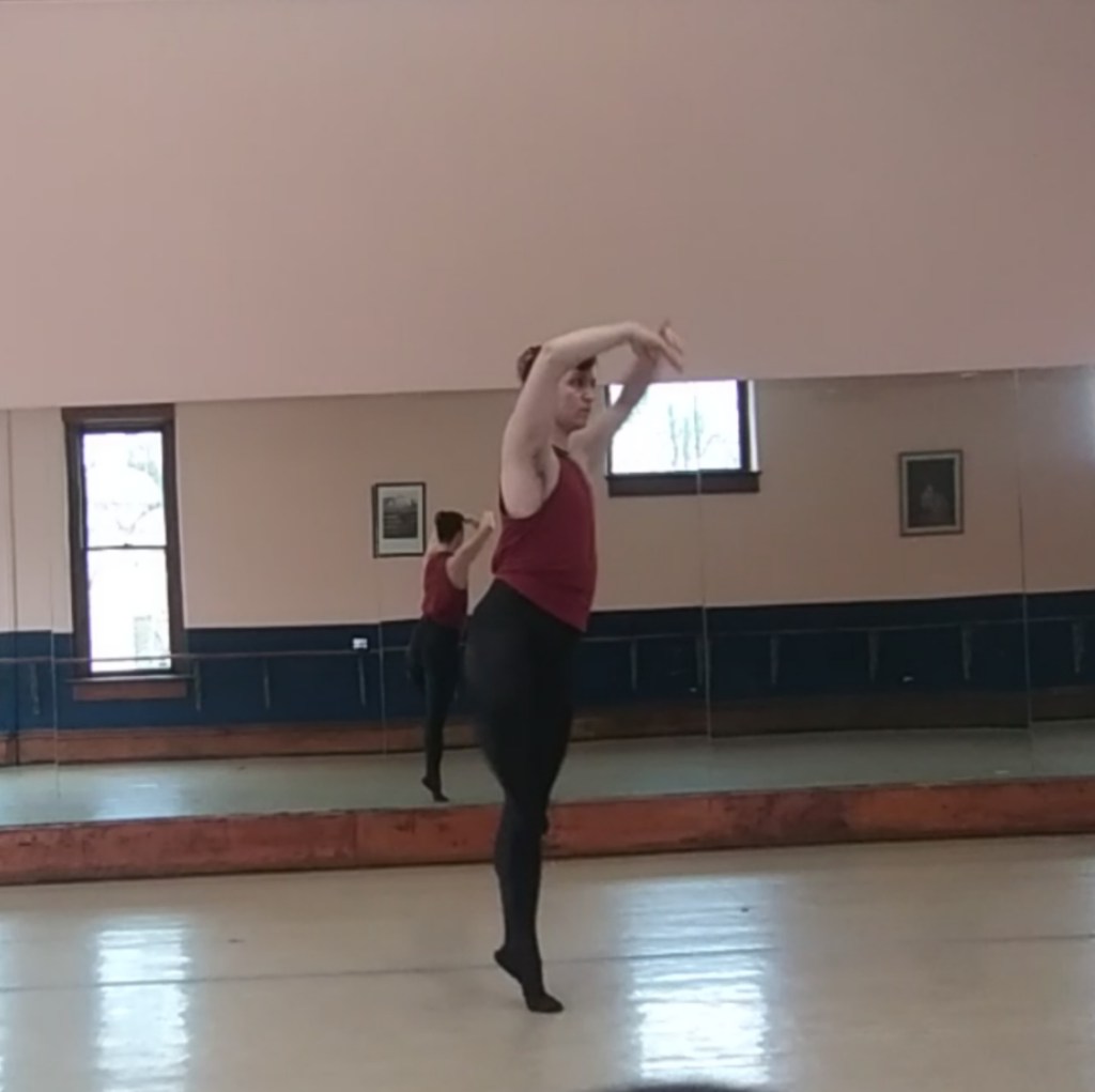 A male dancer (the author) in a burgundy sleeveless shirt, black tights, and black shoes, halfway through a pirouette with his arms curved and raised so his hands are just above eye level, his right leg raised and bent so the toes are touching the bottom of his left knee. He does not appear to be enjoying himself, although as the author and the dancer I can attest that he was.