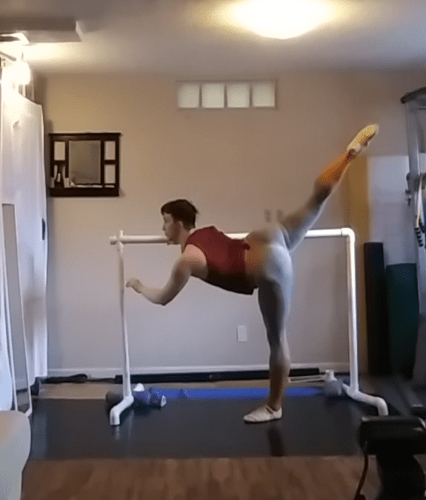 The author at a white pvc ballet barre performing a penche, a tilted balance, with the right leg at 1:00 and the left leg standing at 6:00.